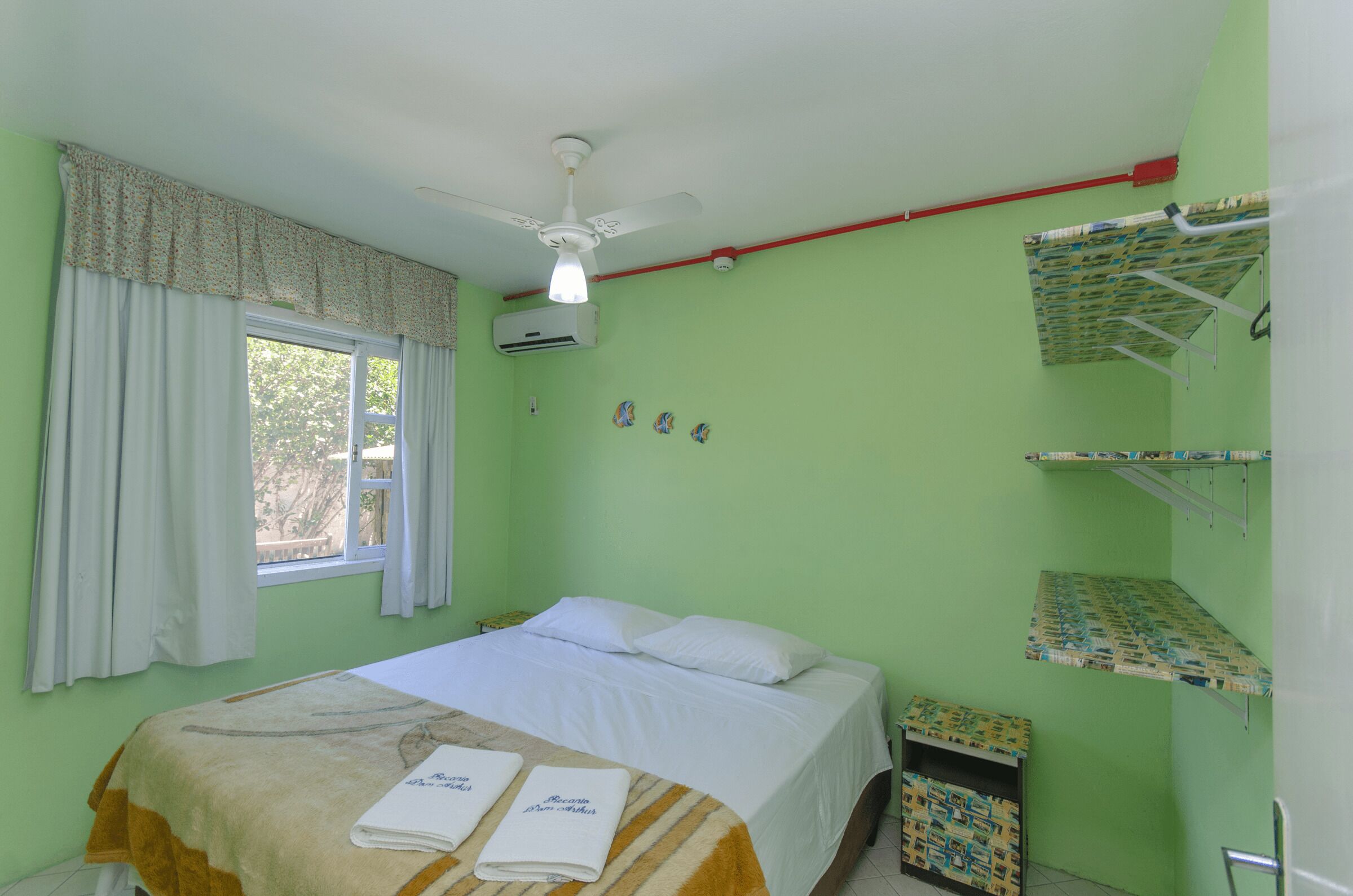 A single bed with white linens, a ceiling fan, an air conditioning unit, a window with curtains, and a small bedside table with a towel.