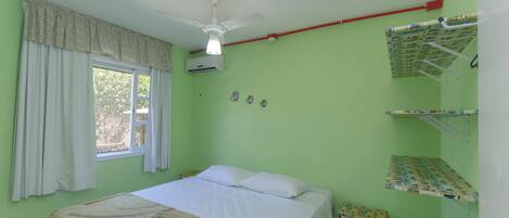 A single bed with white linens, a ceiling fan, an air conditioning unit, a window with curtains, and a small bedside table with a towel.