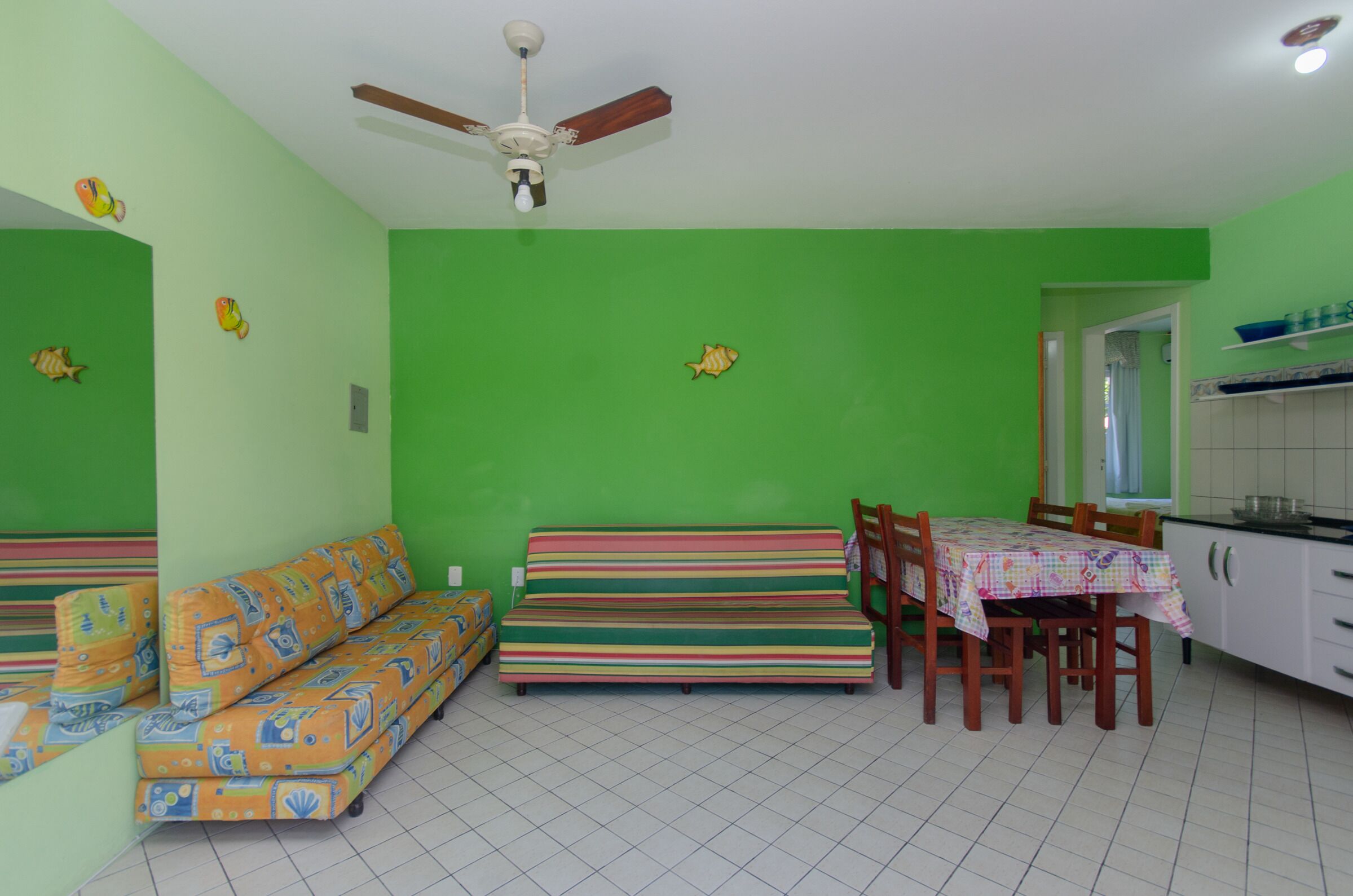 Een kamer met een groene wand, een plafondventilator, een gestreepte bank, een eettafel met stoelen en een keukenhoek.