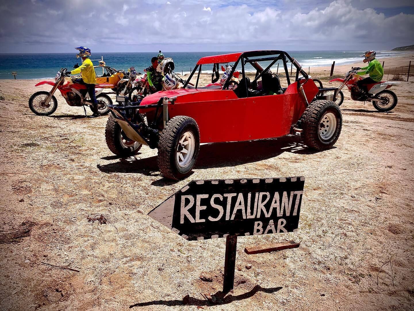 Escena de playa con un vehículo todoterreno rojo estacionado en primer plano y un letrero que dice "RESTAURANT BAR" en el centro.