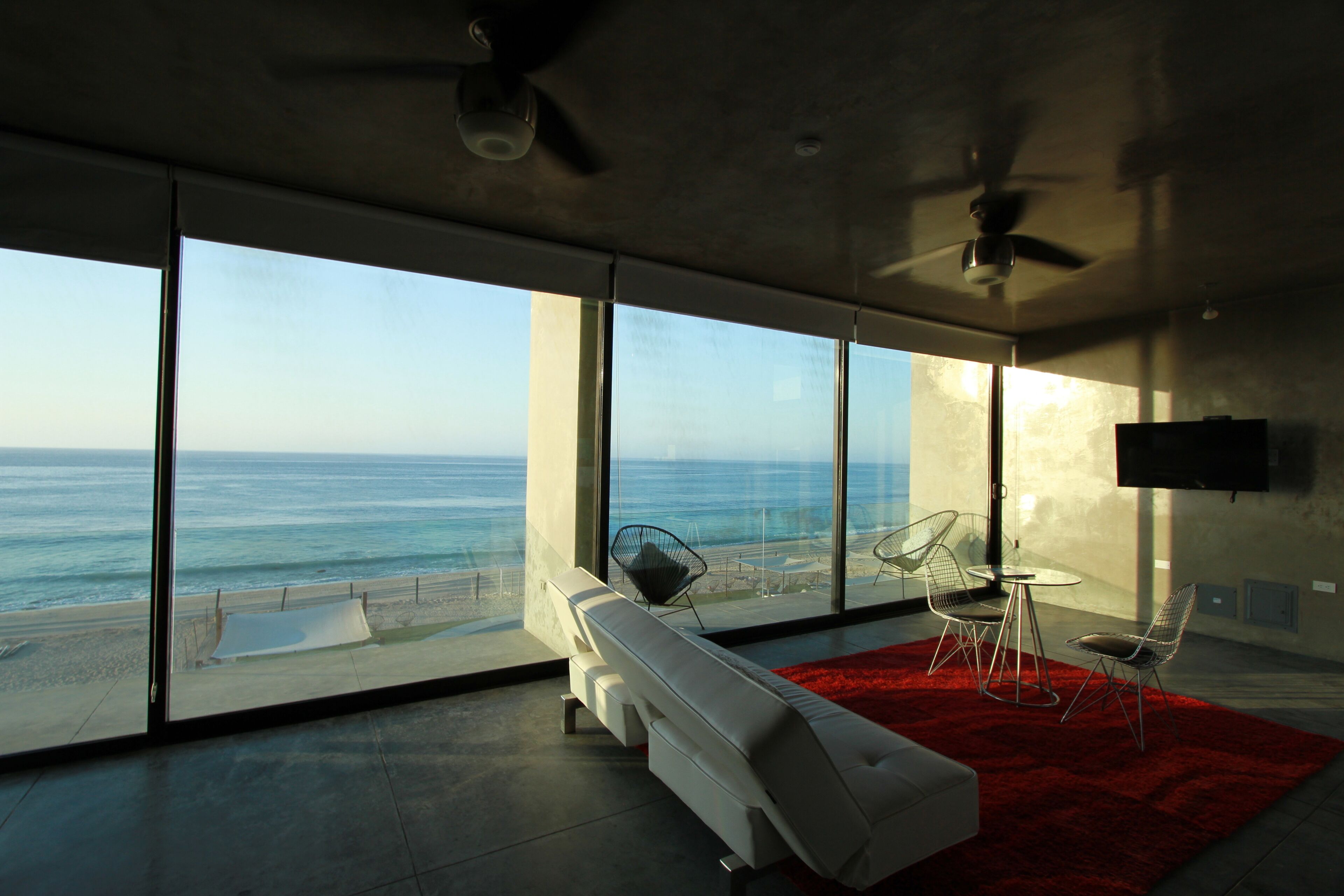 Una habitación moderna con un amplio ventanal con vistas al océano, un sofá blanco, un tapete rojo y un ventilador de techo.