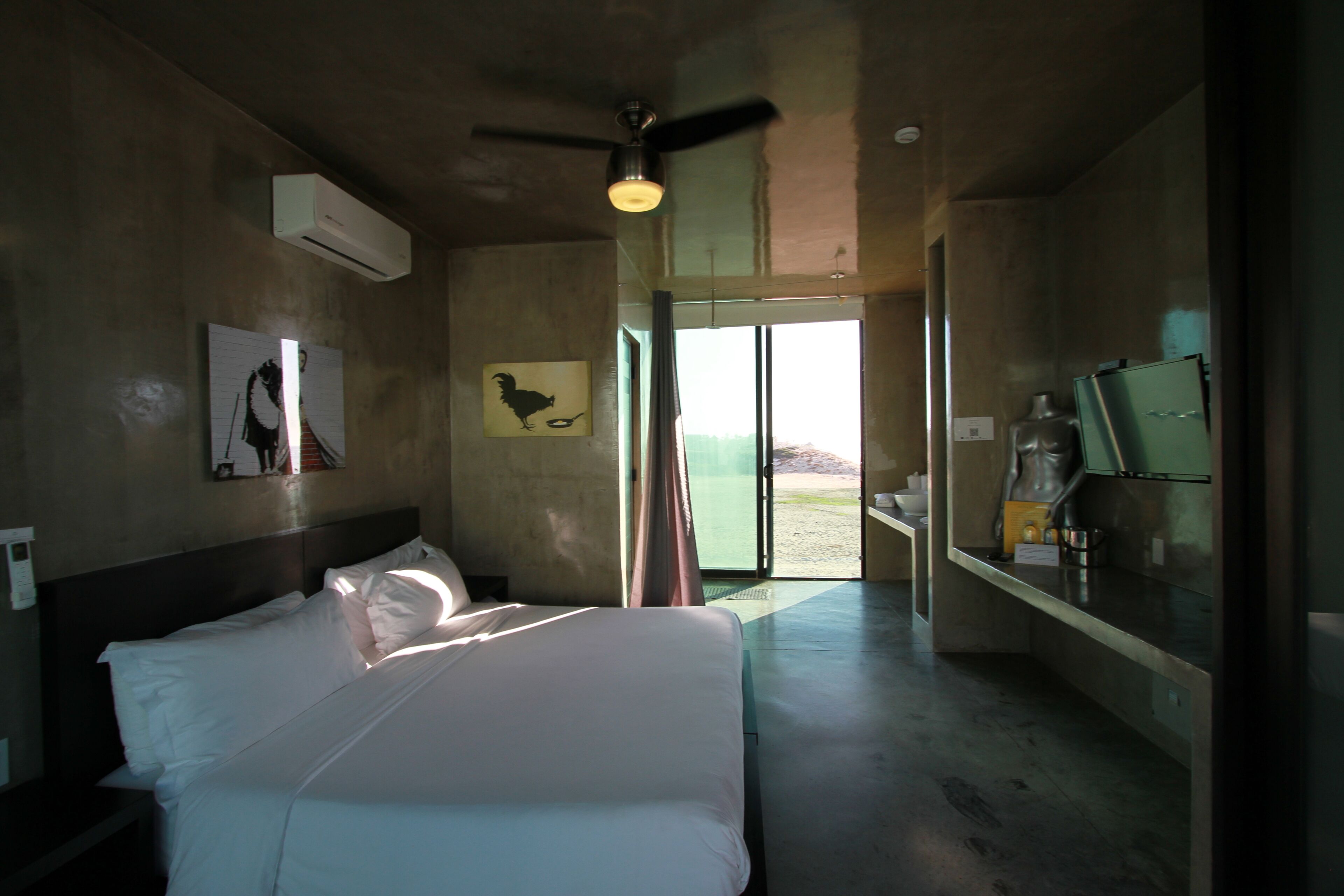 Una habitación de hotel con cama, ventilador de techo, equipo de aire acondicionado, un cuadro, vistas a través de una puerta y un minibar.