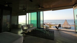 Un dormitorio moderno con una cama grande, una zona de estar con sofá y sillones, y vista al océano a través de ventanales de gran altura.