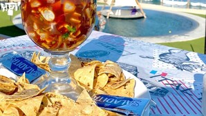 Una bebida tropical con decoración de frutas, totopos y vista a la playa, con un barco al fondo.