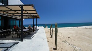 Un área de comedor al aire libre con mesas y sillas, un paseo techado y una playa con cactus.