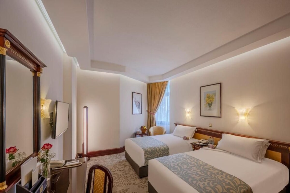 Une chambre d’hôtel avec deux lits, un grand miroir, un bureau avec un téléphone et un vase rempli de fleurs.