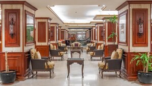 Een ruime lobby met een centrale trap, omringd door rijen fauteuils en tafels, en grote wandlampen.