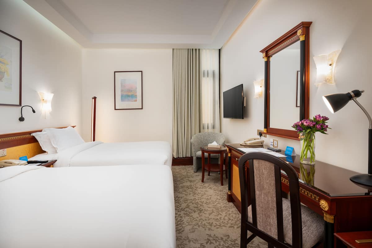 Une chambre d’hôtel avec deux lits, un bureau, une chaise, une lampe de bureau, un vase avec des fleurs, un téléviseur à écran plat, une lampe fixée au mur et un tableau encadré accroché au mur.