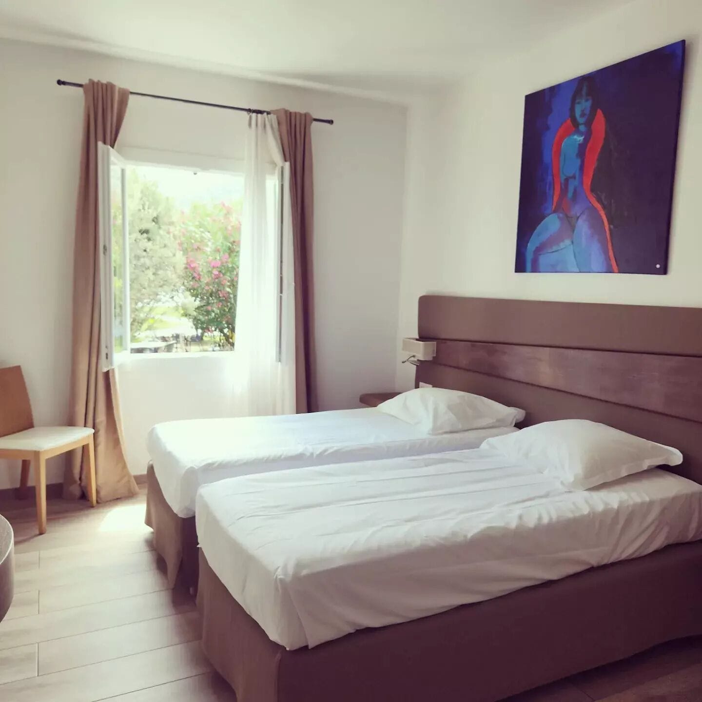 A hotel room with two beds, a window with curtains, a chair, and a painting on the wall.