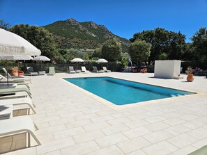 Un área de piscina con sillas de descanso y sombrillas, rodeada de follaje y montañas.
