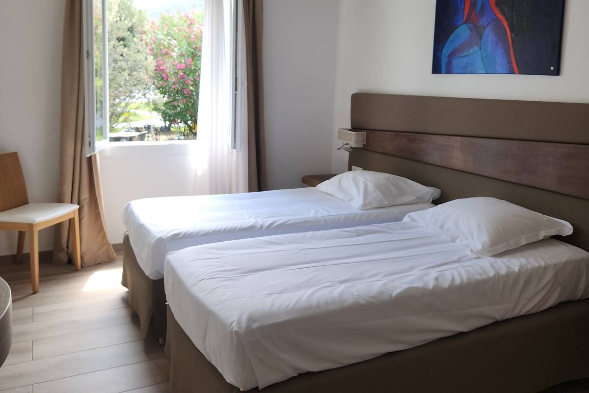 Ein Hotelzimmer mit zwei Betten, Holzboden, einem Fenster mit Vorhängen, einem Gemälde an der Wand und einem Sessel.