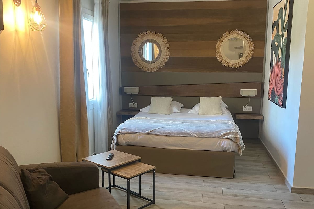 Ein Schlafzimmer mit einem Bett, Nachttischen, einem kleinen Tisch mit Fernbedienung, einem braunen Sofa und einem großen Spiegel an der Wand.