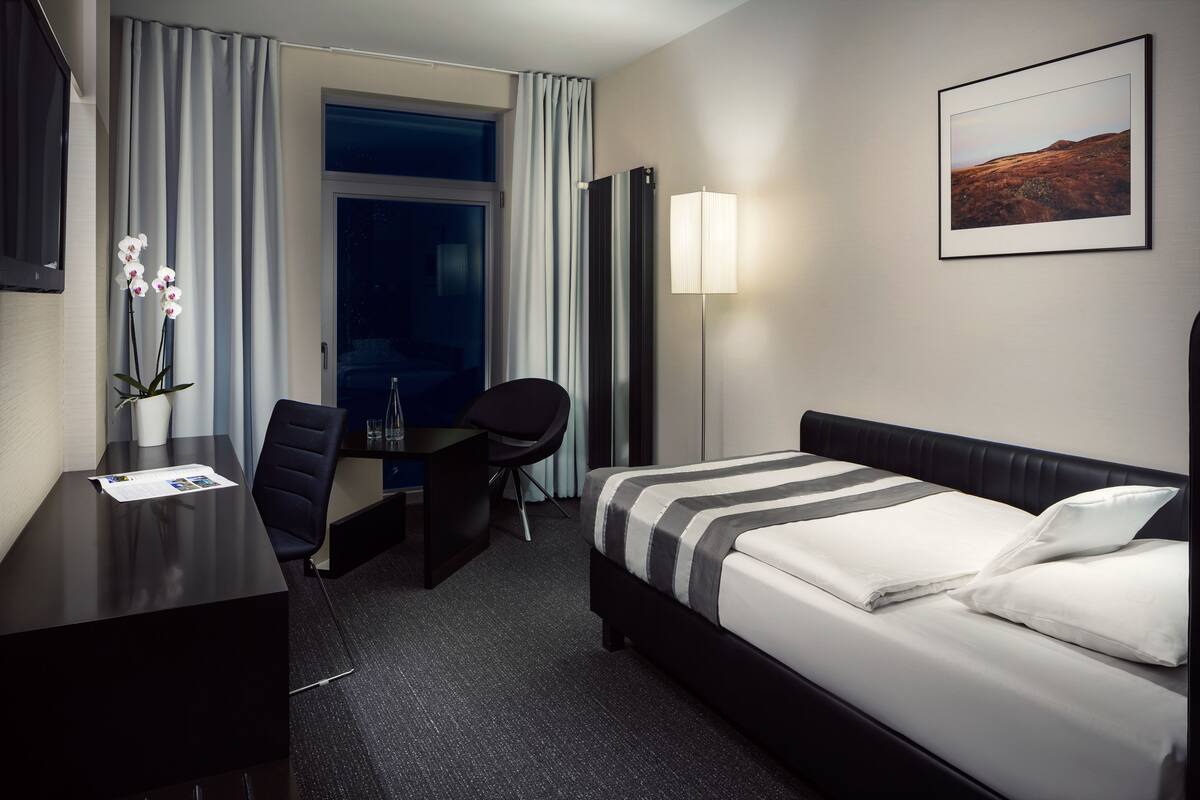 A hotel room with a bed, desk, chair, and a large window.