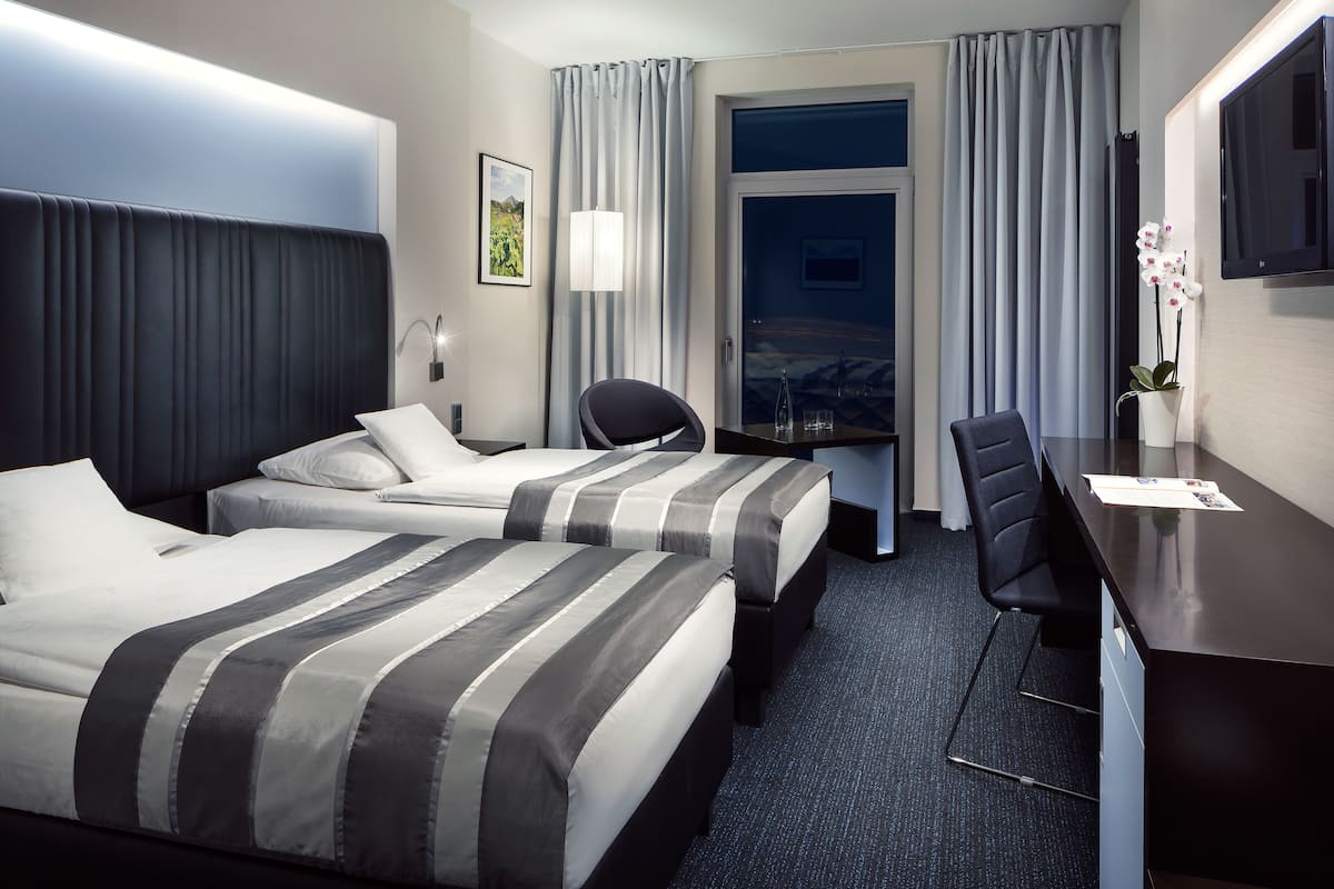 A hotel room with two beds, a desk, a chair, a TV, and a window with curtains.