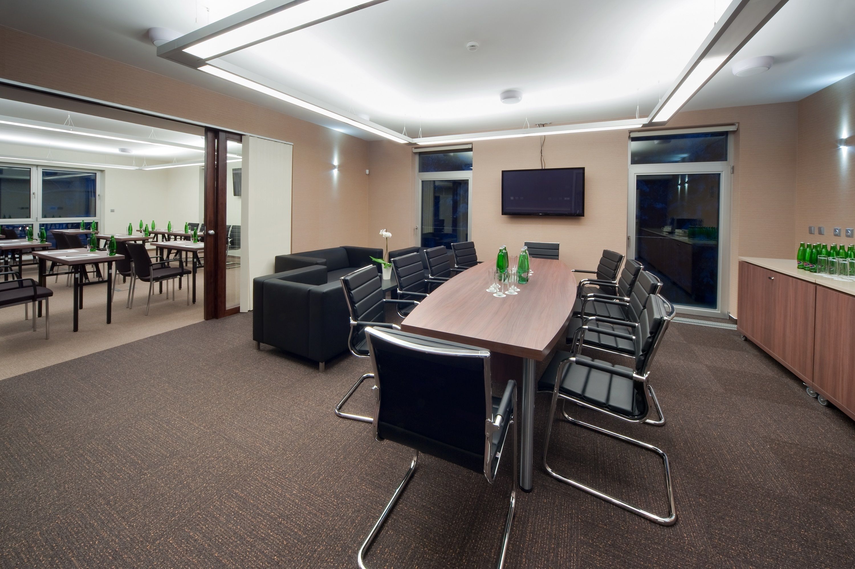 A modern conference room with a long table, black chairs, and a sofa. There are green bottles on the table and a TV on the wall.
