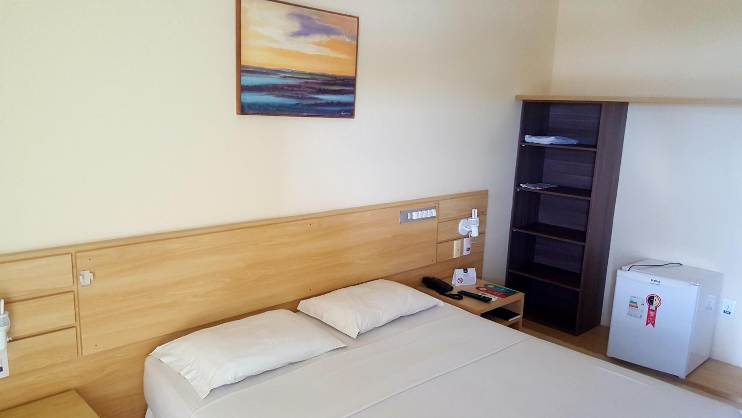 A hotel room with a bed, a nightstand, a small refrigerator, a wardrobe, and a painting on the wall.