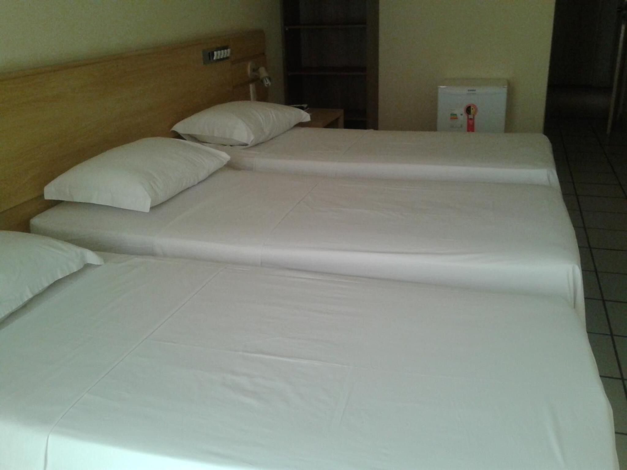 Quarto de hotel com três camas de solteiro, cada uma com roupa de cama e almofadas brancas. Há uma cabeceira de madeira, uma mesinha com abajur e um frigorífico no canto.