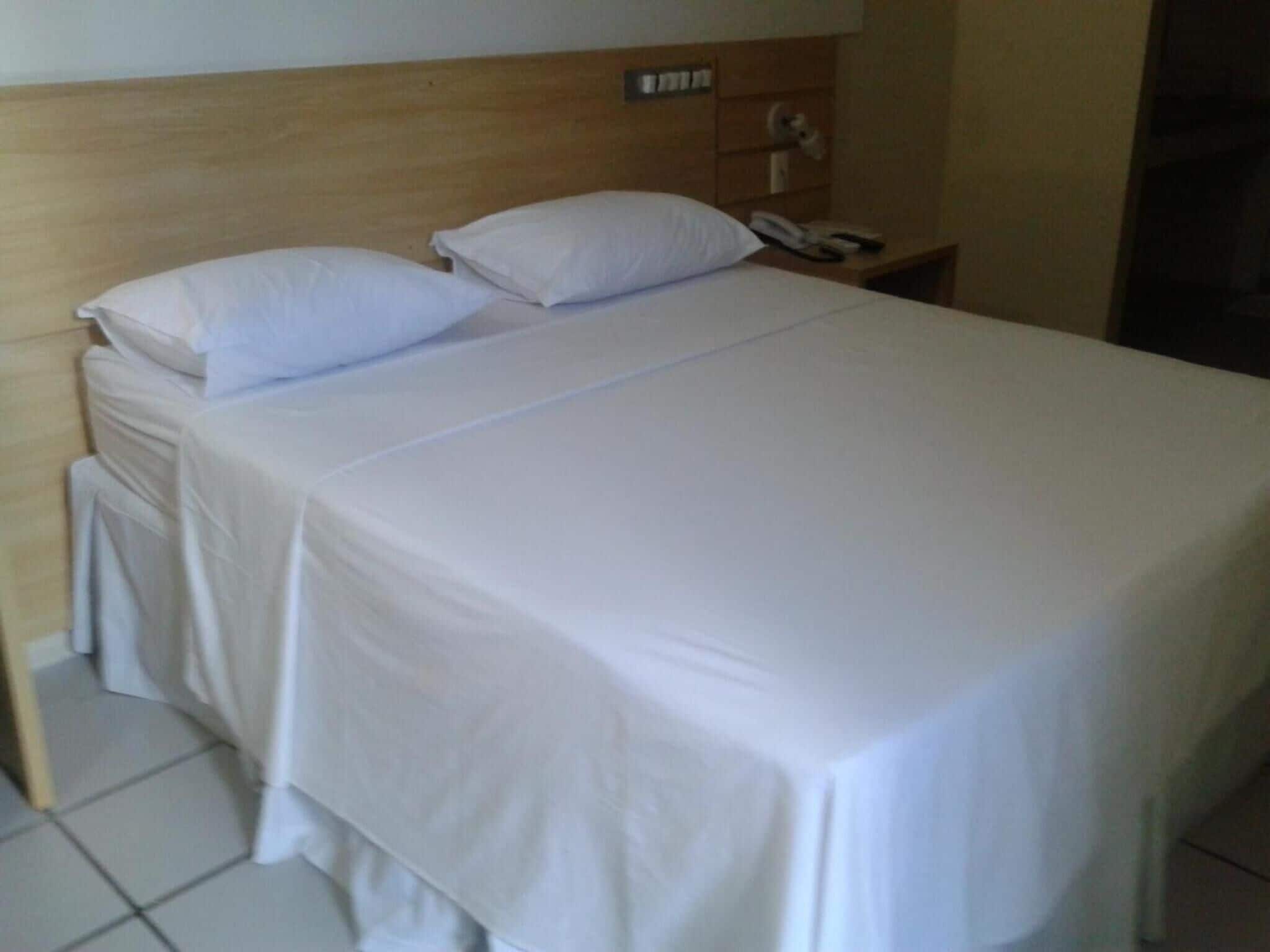 Una cama bien hecha con sábanas blancas, cabecera de madera y mesita de noche con un teléfono.