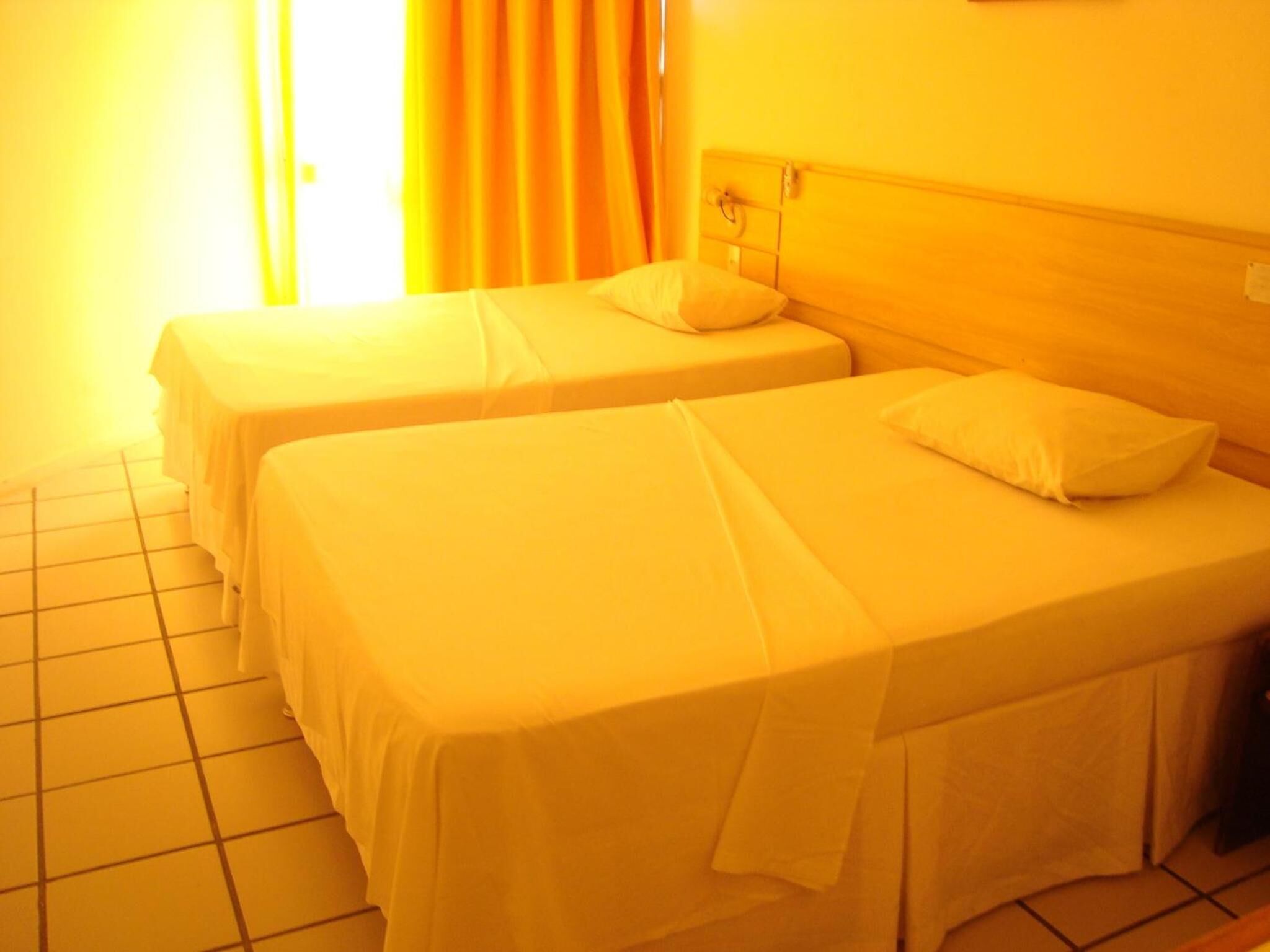 Quarto de hotel com duas camas, piso ladrilhado e janela com cortinas.