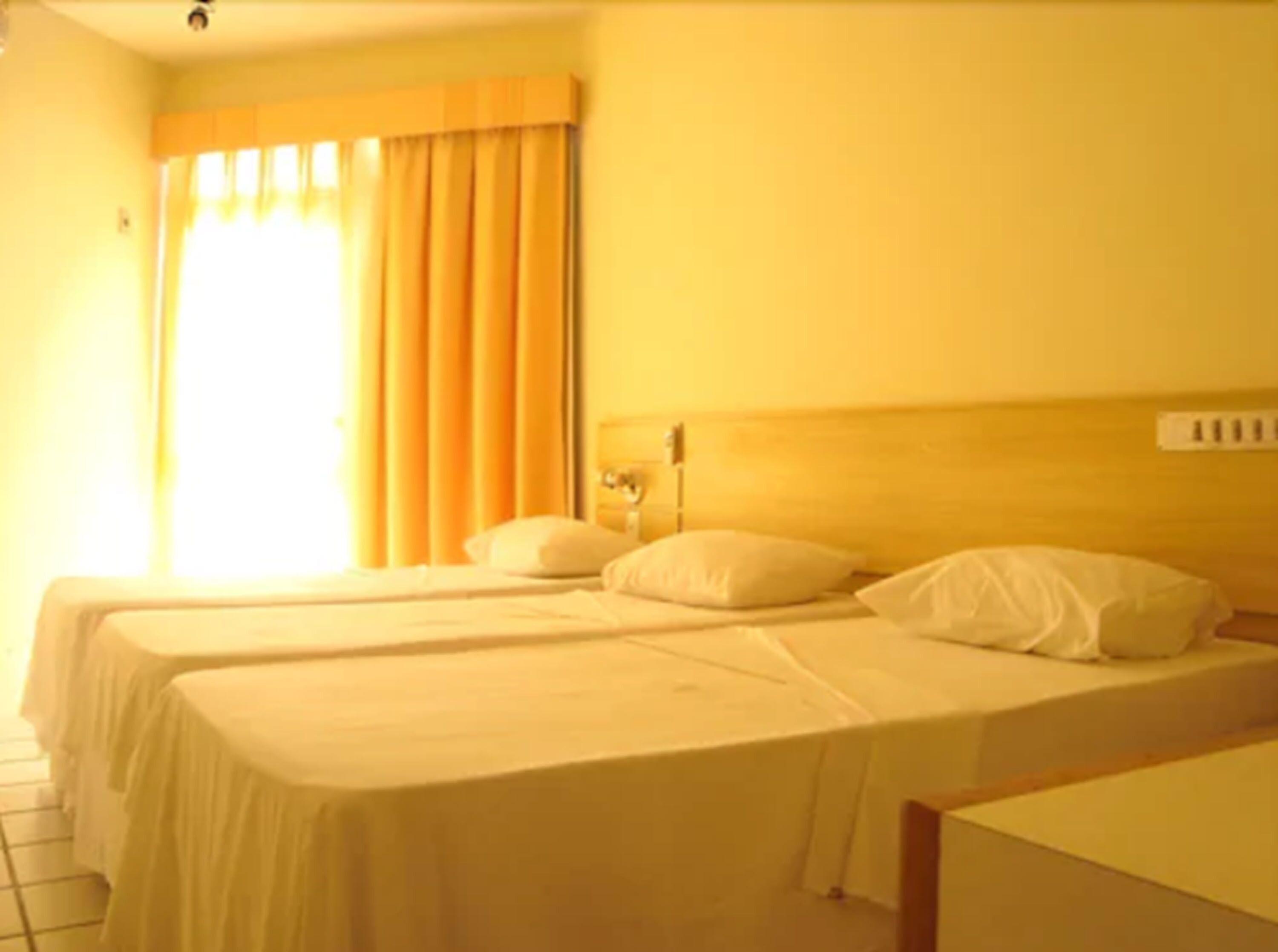 Camera d'albergo con due letti singoli, una testiera in legno, una finestra con tende e un portalampada a parete.
