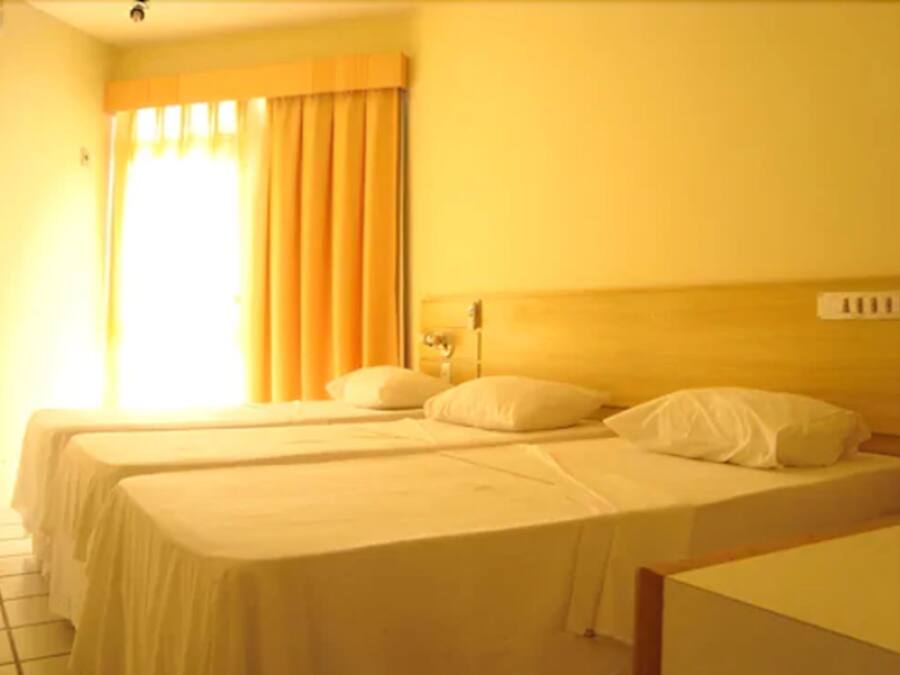 Habitación de hotel con dos camas individuales, cabecero de madera, ventana con cortinas e iluminación montada en la pared.