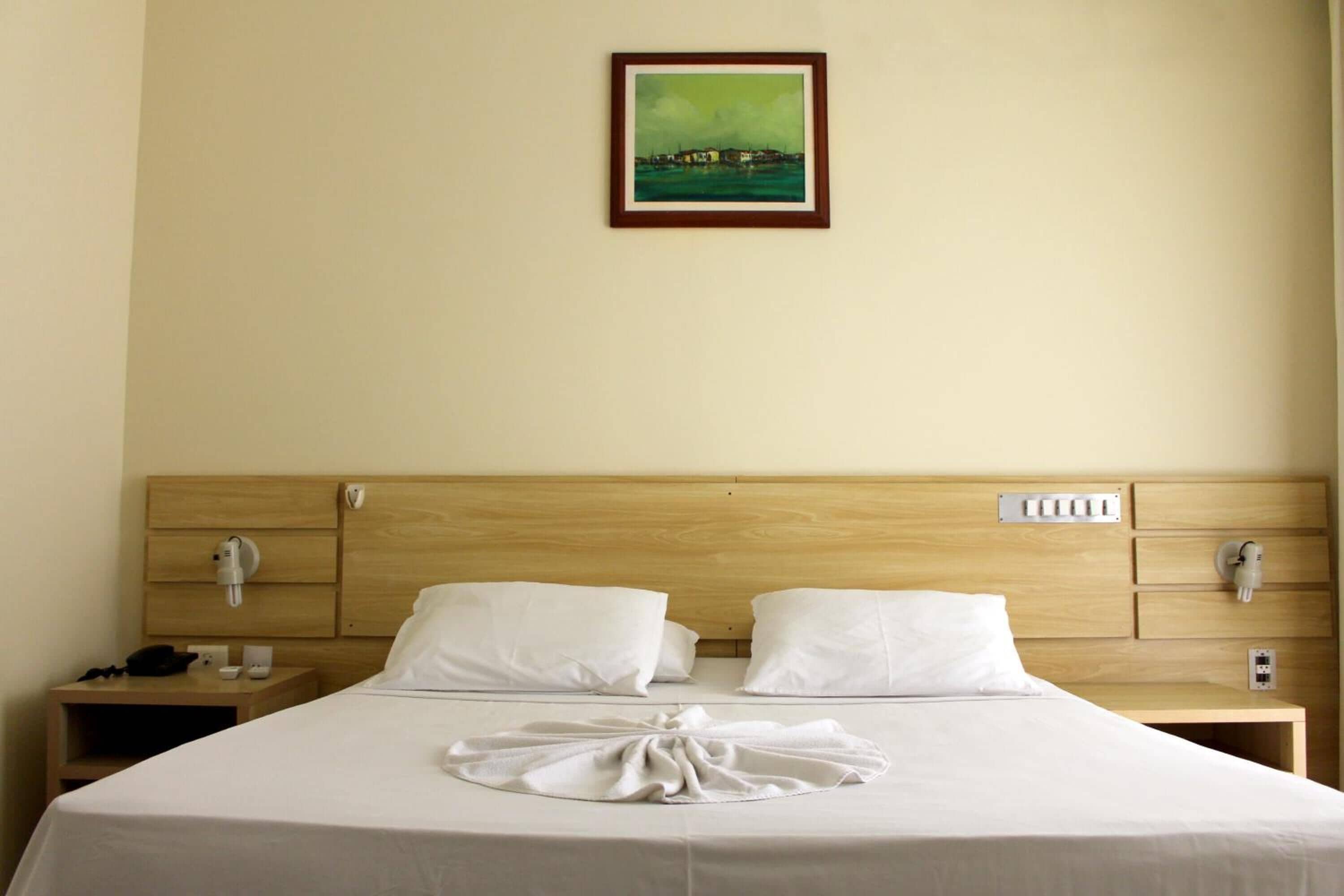 Quarto de hotel com cabeceira de madeira, roupa de cama branca, cama bem arrumada com toalha, mesa de cabeceira com abajur e um quadro na parede.