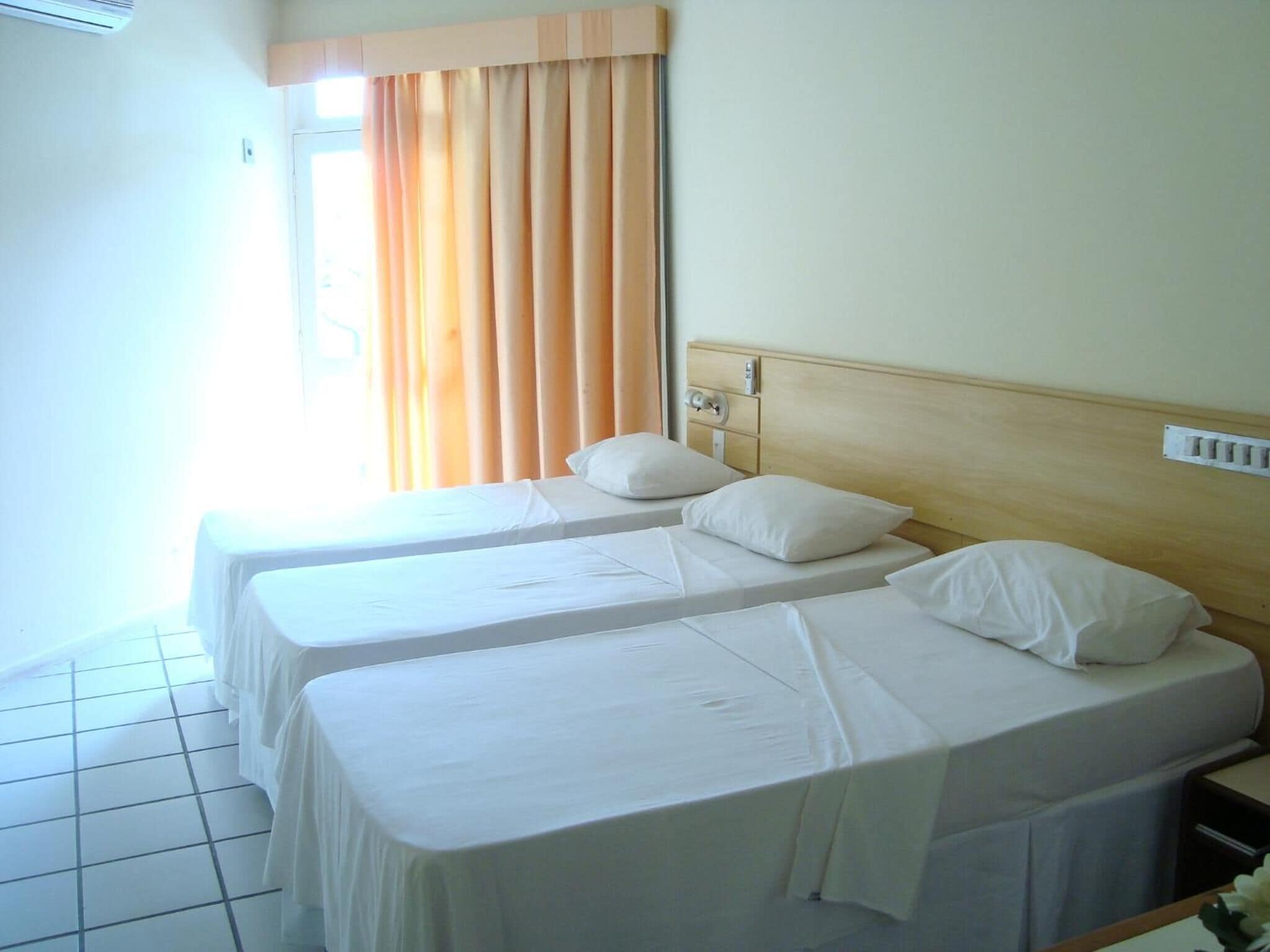 Una habitación de hotel con dos camas individuales, cabecero de madera, ventana con cortinas y suelo de baldosas.