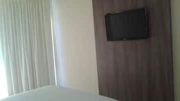 A hotel room with a bed, a television mounted on a wooden panel, and a window with curtains.