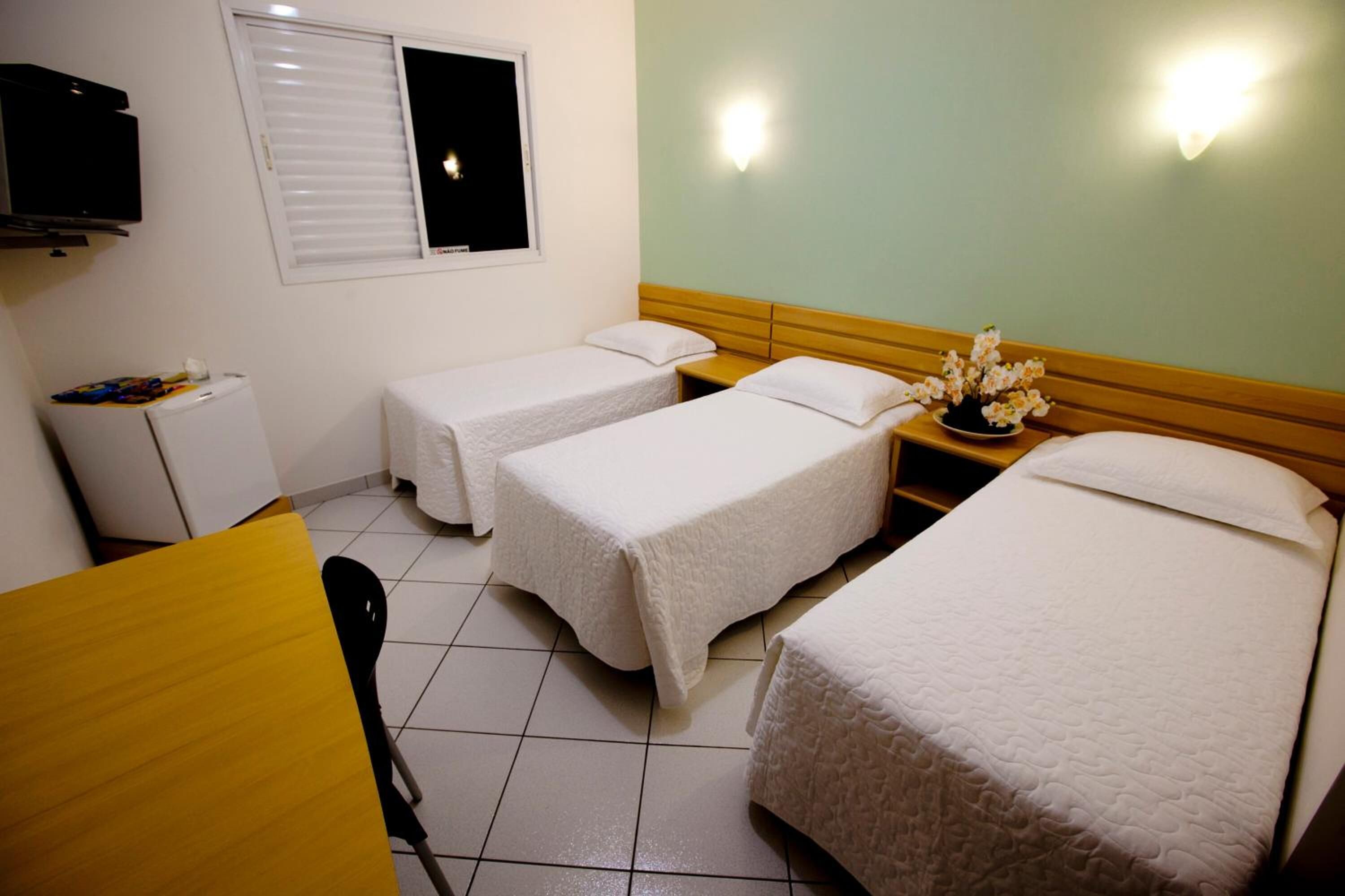 A hotel room with two single beds, a small table, a chair, a television, and a window with blinds.
