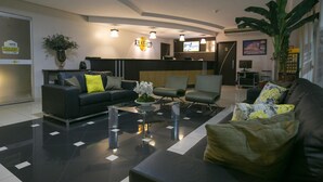 Een moderne hotellobby met een zwart-wit betegelde vloer, een receptiebalie, een bank met kussens, een salontafel en een grote plant.