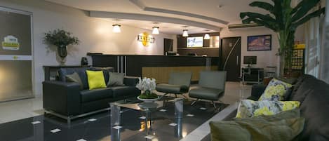 Un moderno vestíbulo de hotel con piso de baldosas blanco y negro, una recepción, un sofá con cojines, una mesa de centro y una planta grande.