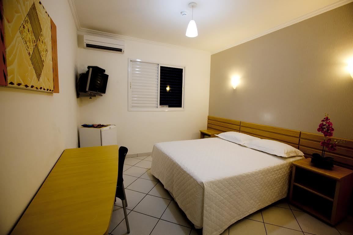 Ein Hotelzimmer mit Bett, Schreibtisch, Stuhl, einem kleinen Tisch, einem an der Wand montierten Fernseher und einem Fenster mit Jalousien.