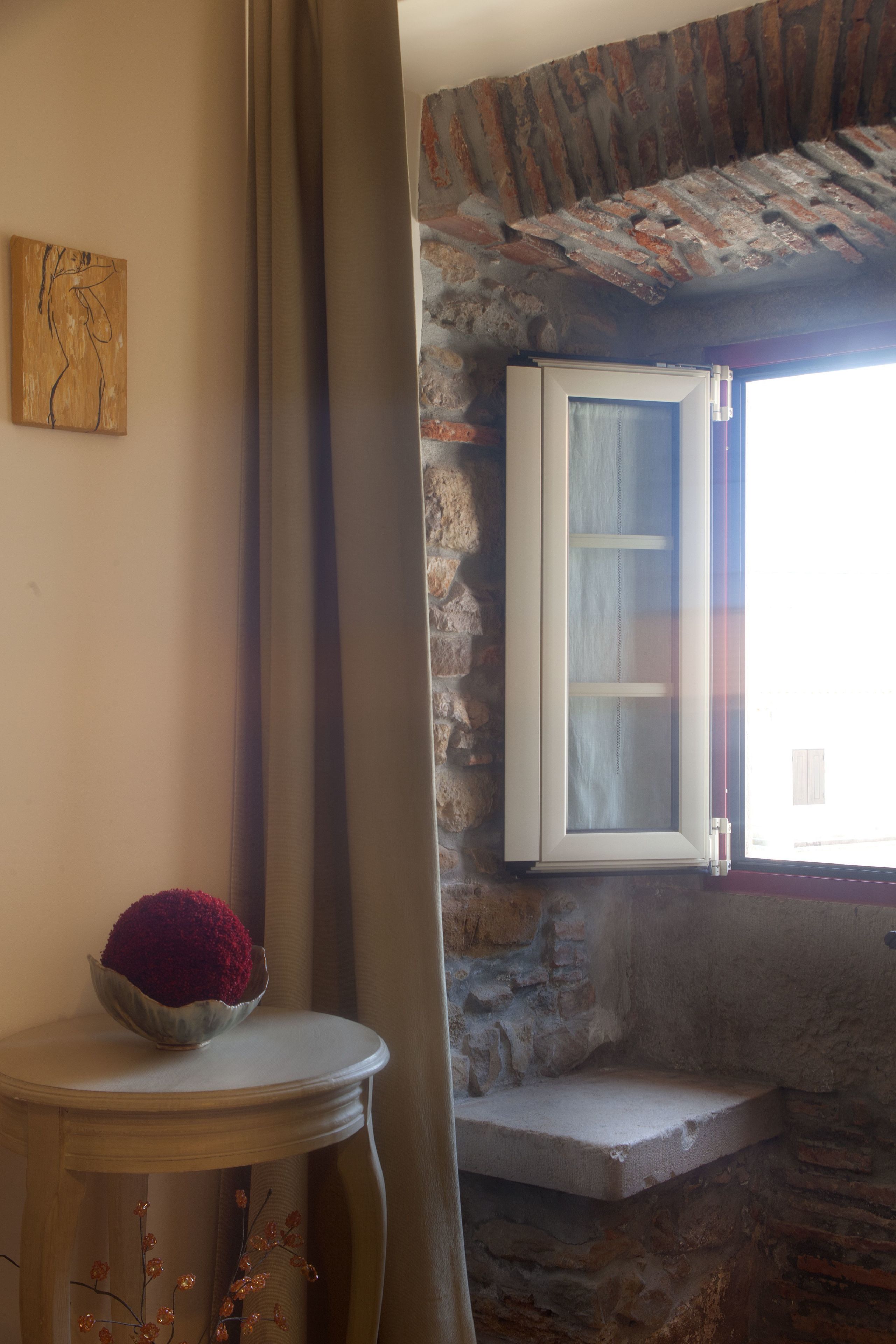Una habitación con pared de piedra, ventana con marco blanco, una mesita con un cuenco de flores rojas y una obra de arte abstracta en la pared.
