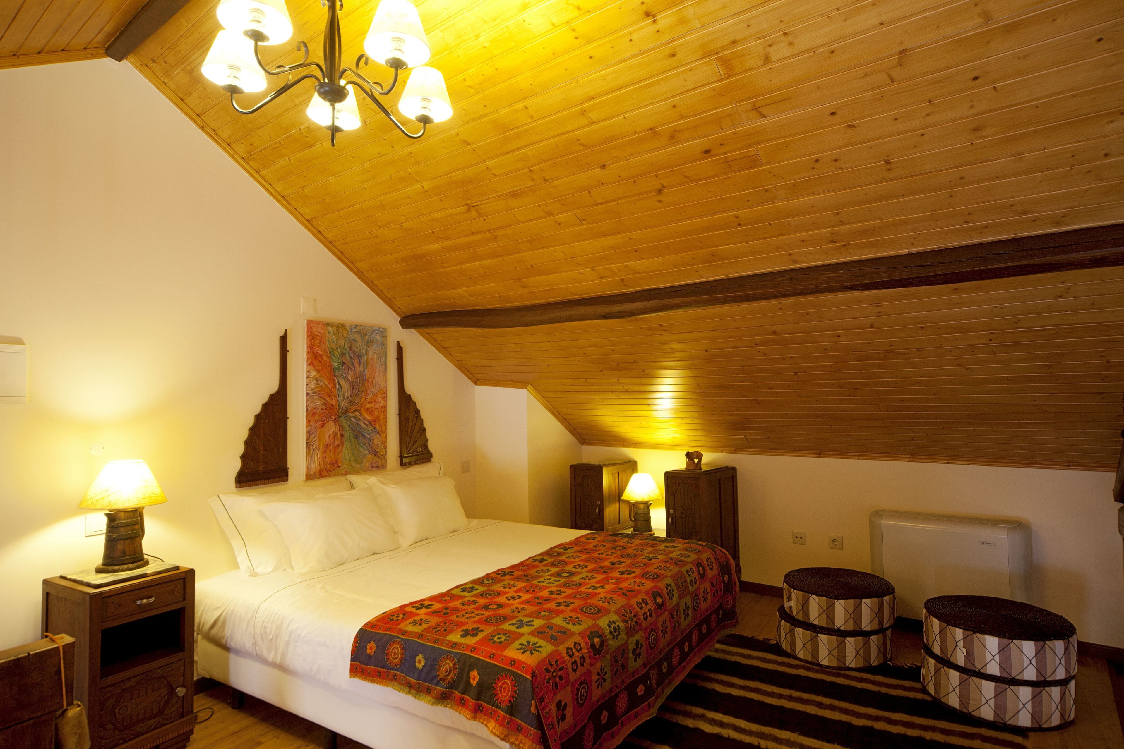 Un dormitorio acogedor con techo de madera, una cama con manta colorida, dos lámparas de noche y un radiador.