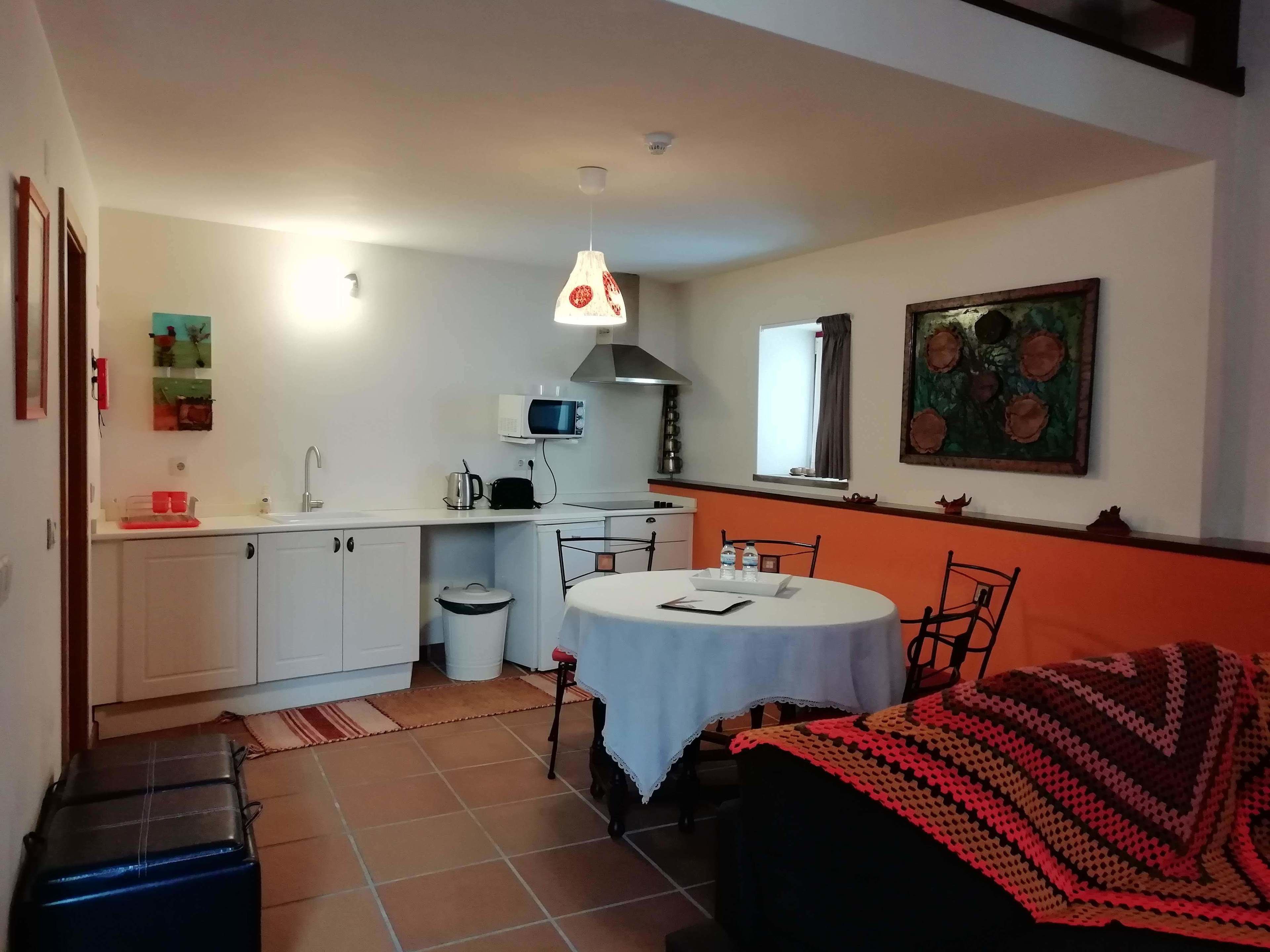 Cocina compacta con armarios blancos, microondas y lavamanos. Hay una mesa de comedor con sillas y una manta.