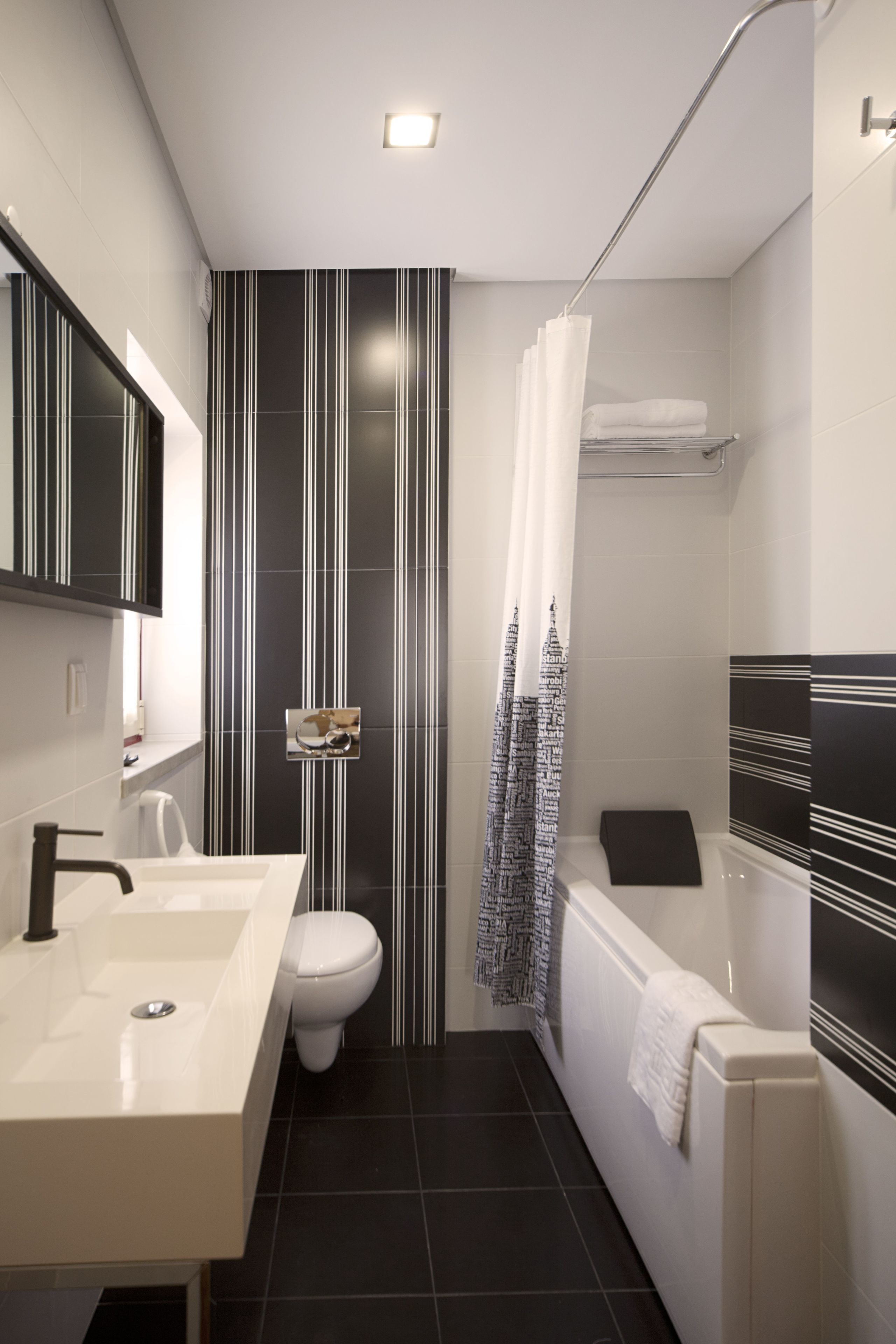 Een moderne badkamer met een wit wastafel, toilet en bad. De wand is gestreept en er is een douchegordijn met een stadsgezicht dessin.