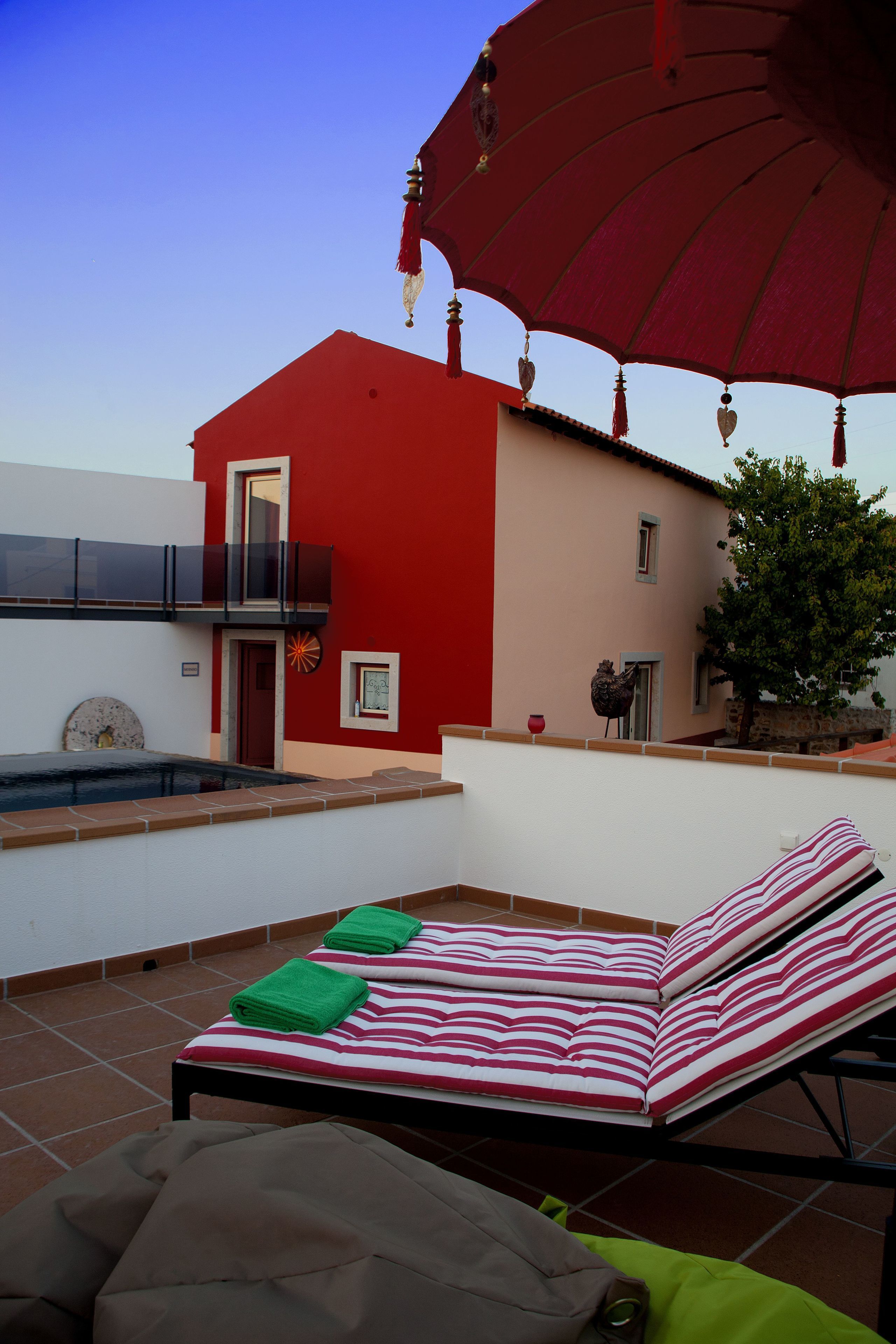 A red and white building with a pool, a red umbrella, and a lounge chair with striped cushions.