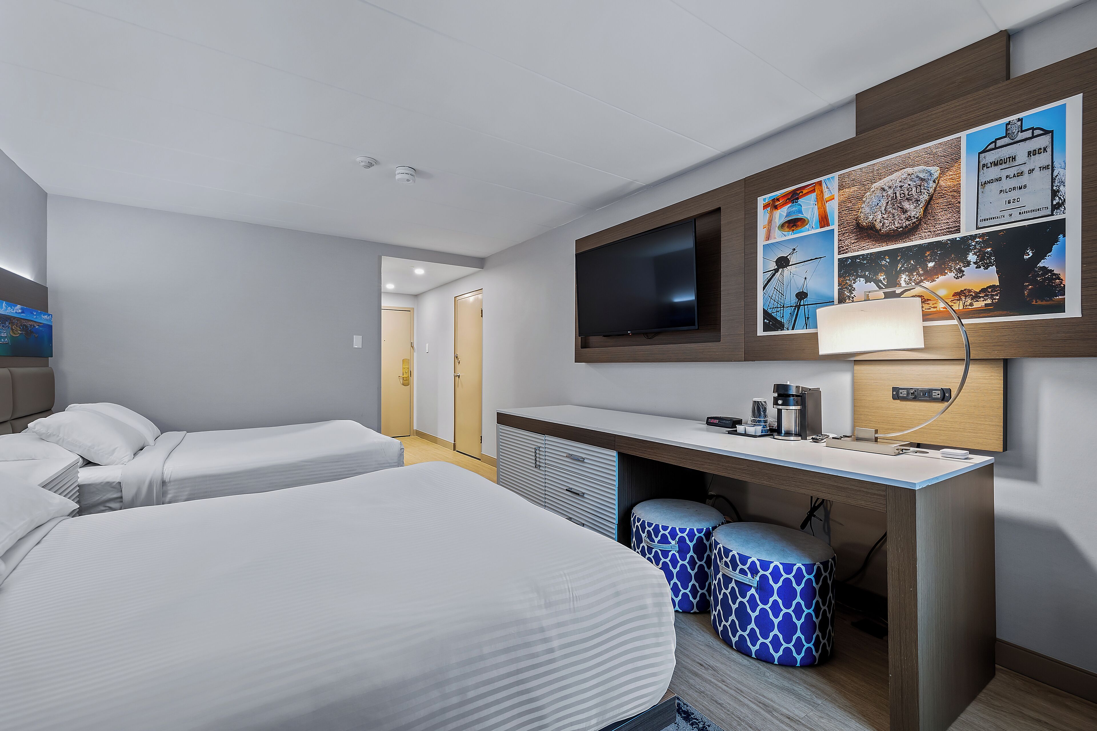 Une chambre d’hôtel avec deux lits, un bureau doté d’un téléviseur à écran plat, une machine à café et un tableau accroché au mur représentant Plymouth, lieu de pèlerinage.