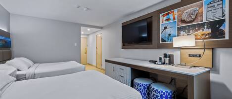 A hotel room with two beds, a desk with a flat-screen TV, a coffee maker, and a wall-mounted picture of Plymouth Place of Pilgrims.