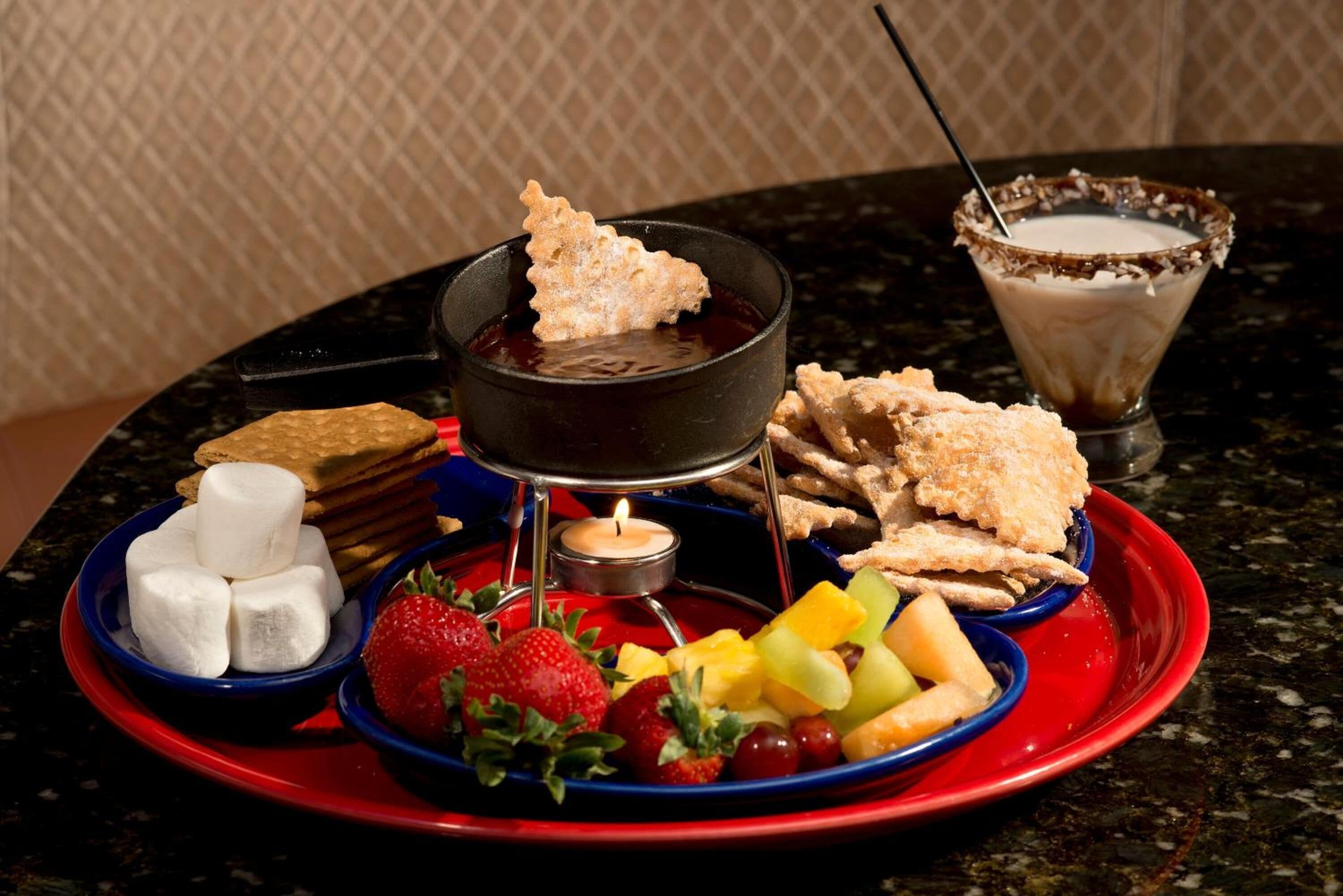 A dessert setup with a fondue pot, fruit, marshmallows, and chocolate-covered cookies on a red plate.