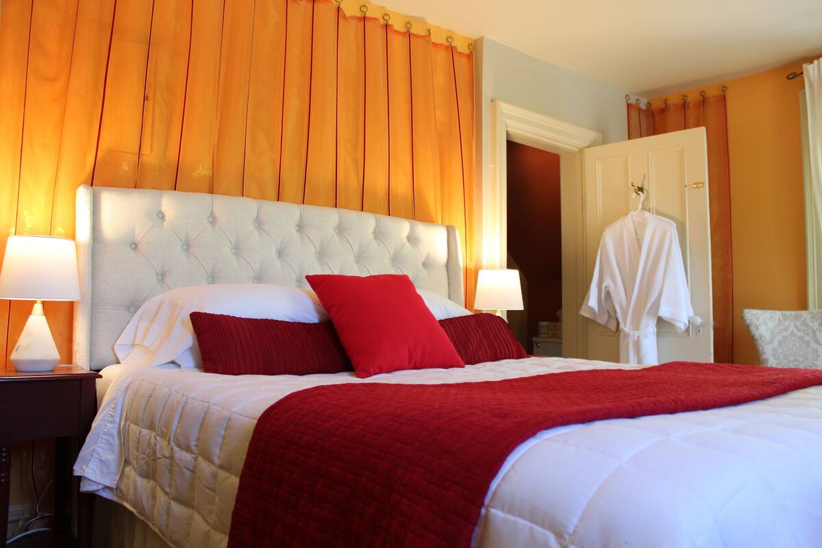 A hotel room with a large bed, two bedside lamps, a red throw blanket, and a white robe hanging on the door.