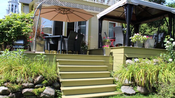 A covered outdoor seating area with steps leading up to a porch, surrounded by greenery and rocks. - Auberge La belle Victorienne (Magog)
