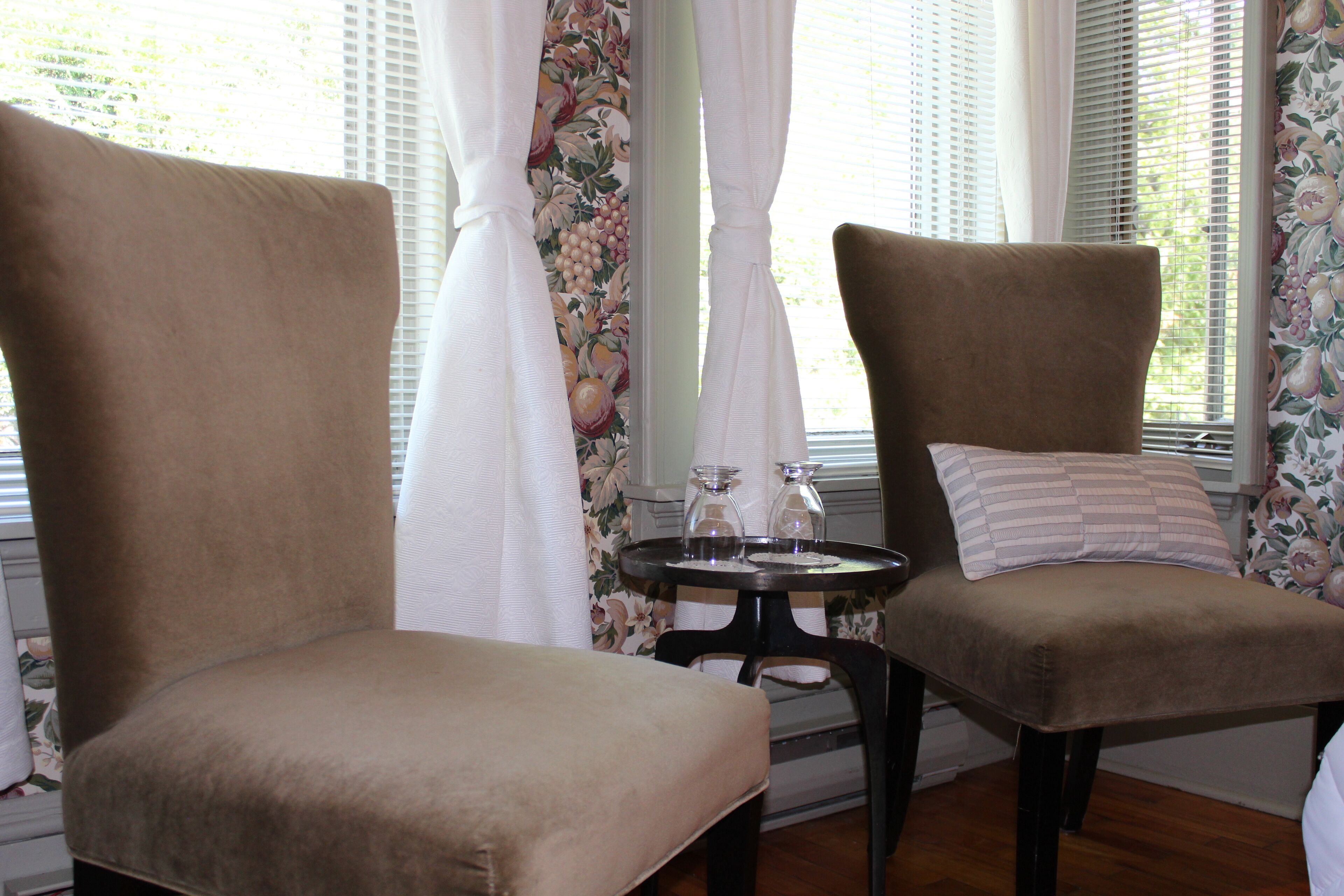 Dos sillones marrones tapizados con una pequeña mesa redonda entre ellos, una cortina blanca y papel tapiz con motivos florales.