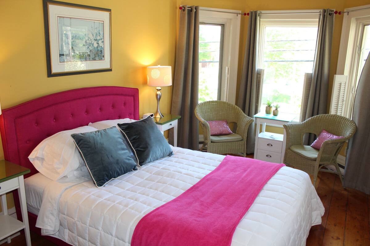 A hotel room with a large bed, a bedside table, a lamp, two chairs, and a window with curtains.