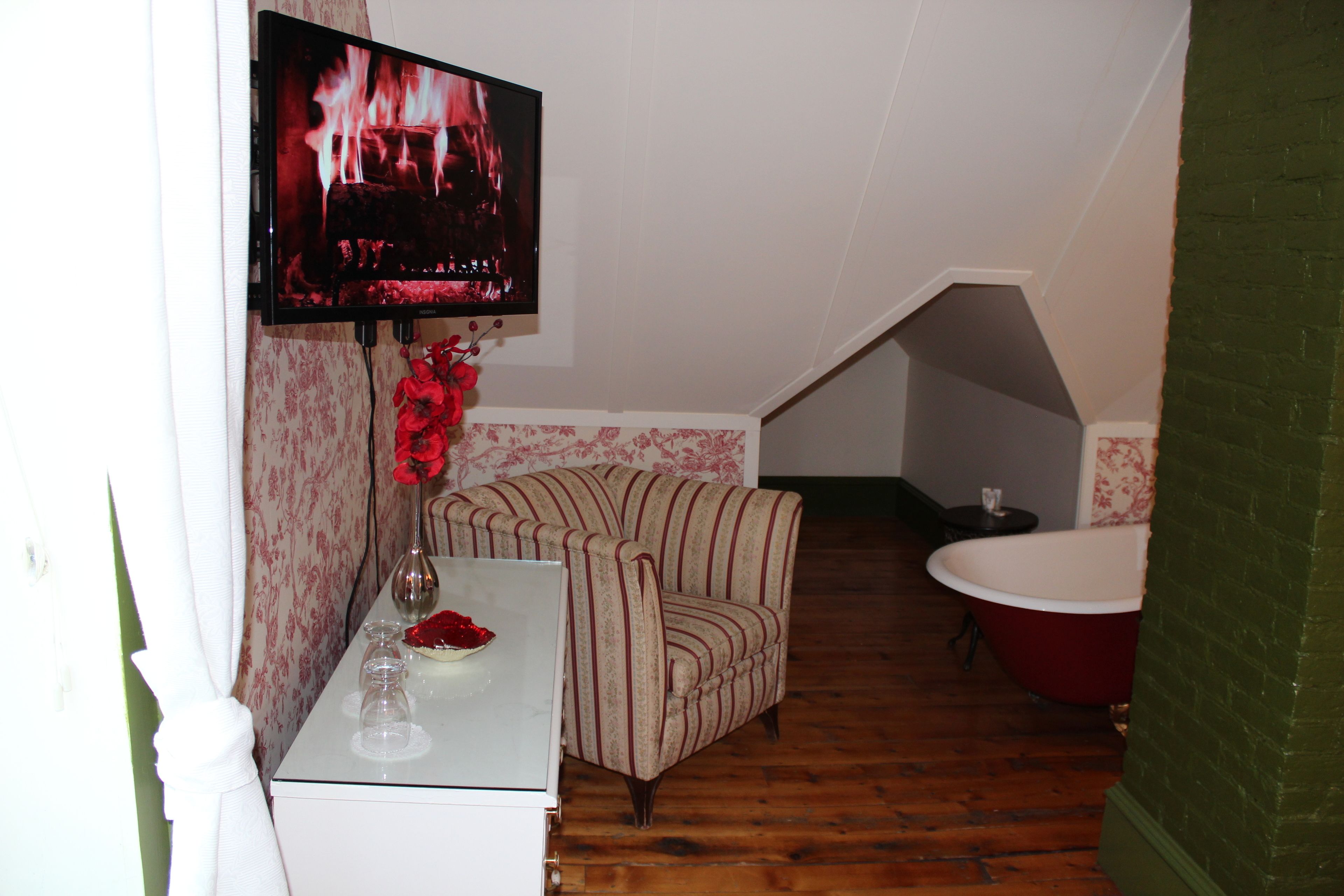 A small, cozy room with a television displaying a fire, a striped armchair, a small table with a vase of red flowers, and a red bathtub.
