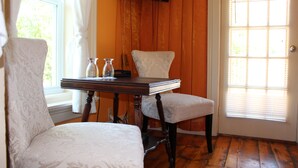 A room with a wooden floor, a white upholstered chair, a matching ottoman, a small wooden table with a book and two glass bottles, and a window with blinds. - Auberge La belle Victorienne (Magog)
