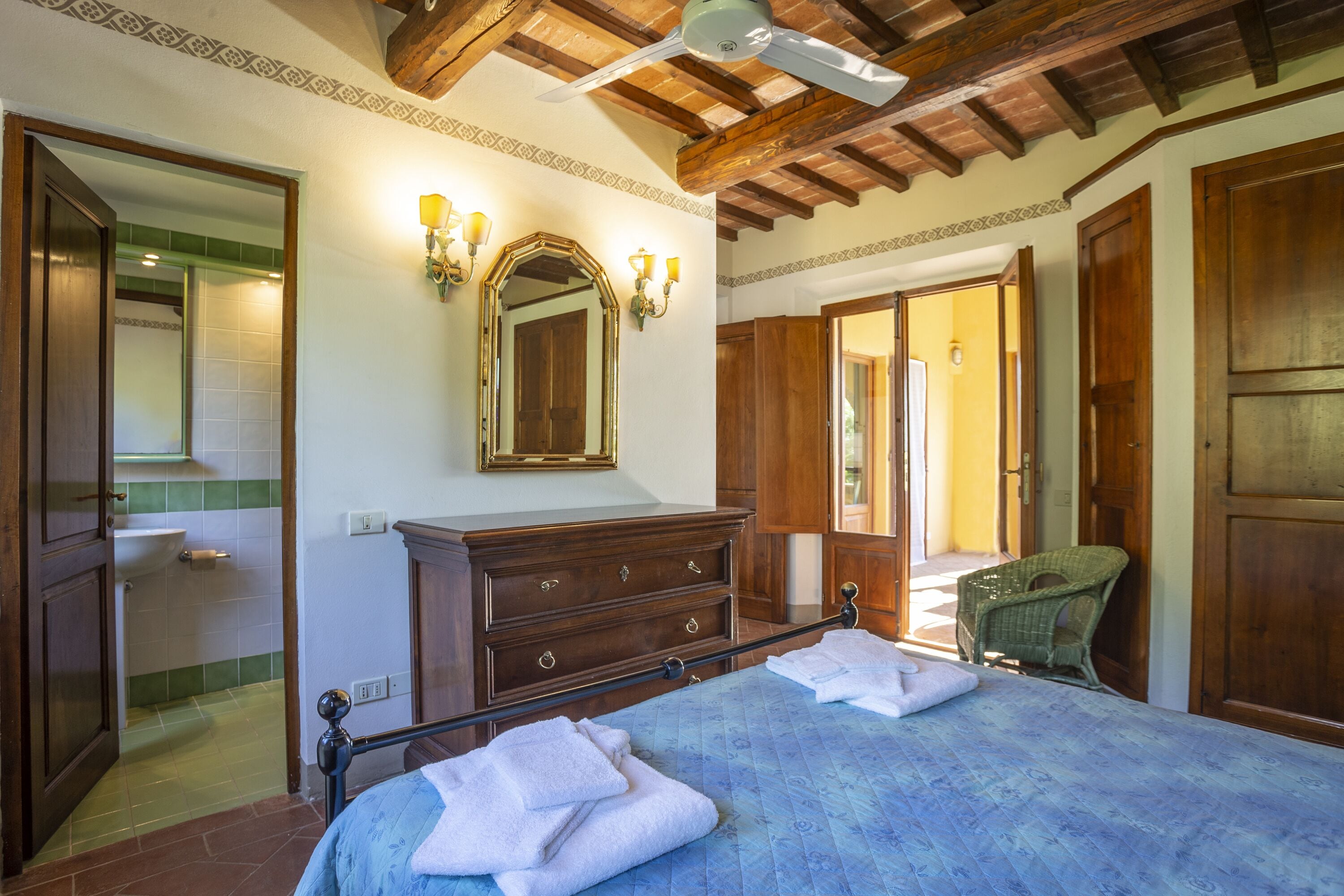 Een slaapkamer met een bed, een houten dressoir en een spiegel. Er staat een stoel en er is een deur naar een andere kamer.
