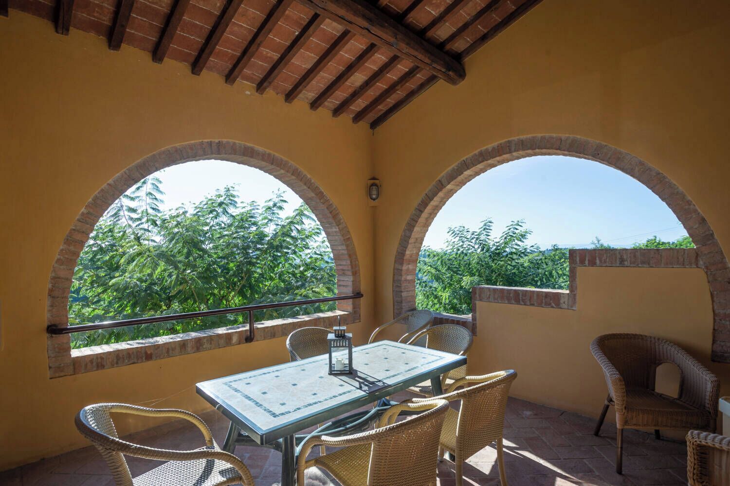 Eine überdachte Terrasse mit Tisch und Stühlen, zwei Rundbogenfenstern und Blick auf Grünflächen.