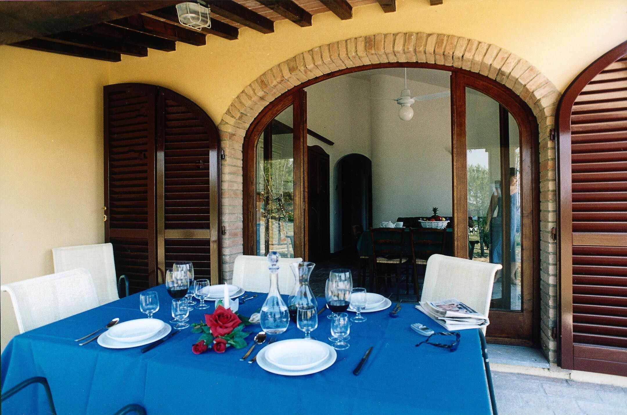 Tavolo apparecchiato per due, con tovaglia blu, piatti bianchi e bicchieri da vino, in una stanza con persiane in legno e un arco in mattoni.