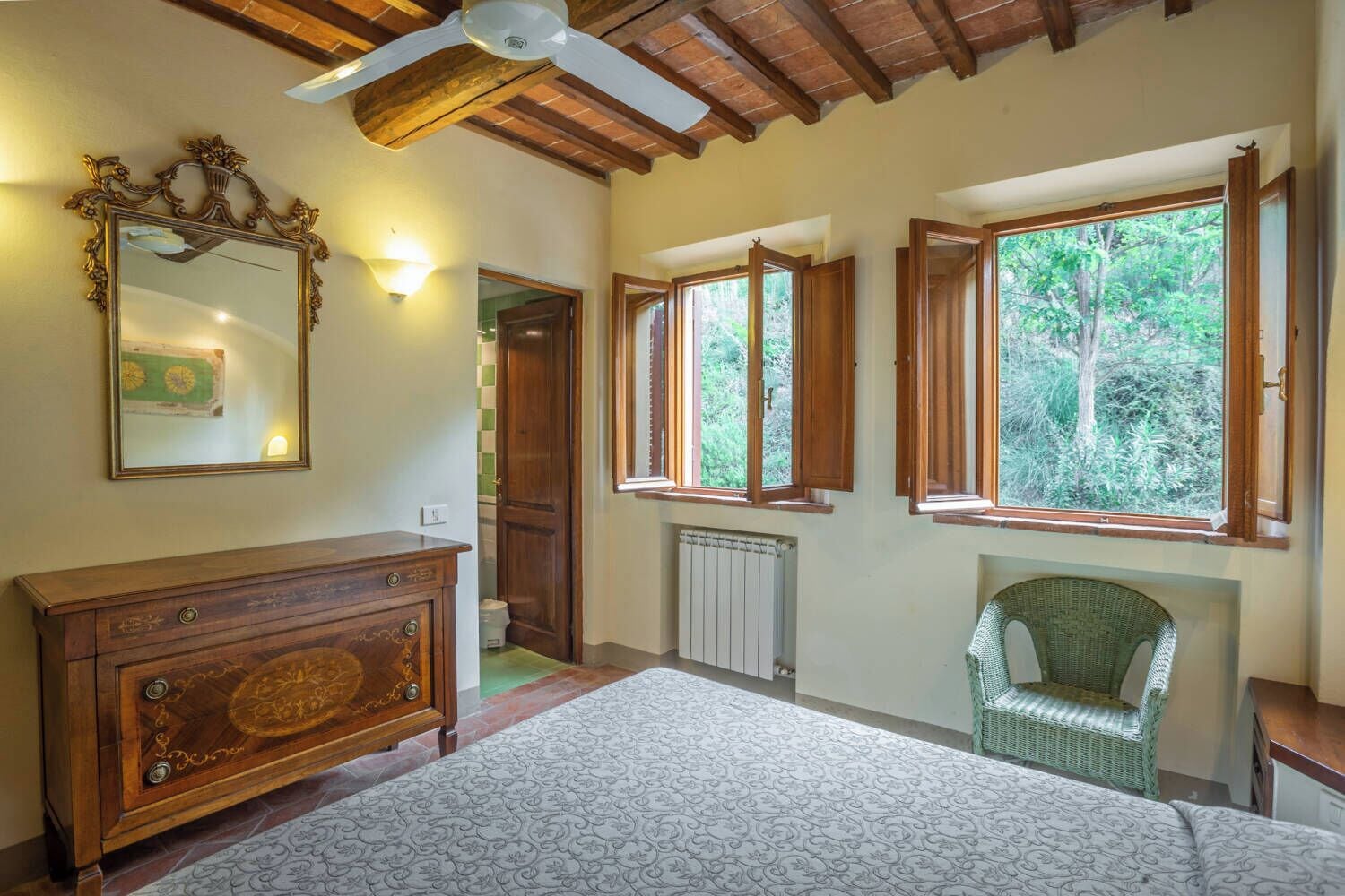A room with a wooden ceiling, a large mirror, a dresser, a radiator, and a window with a view of greenery.